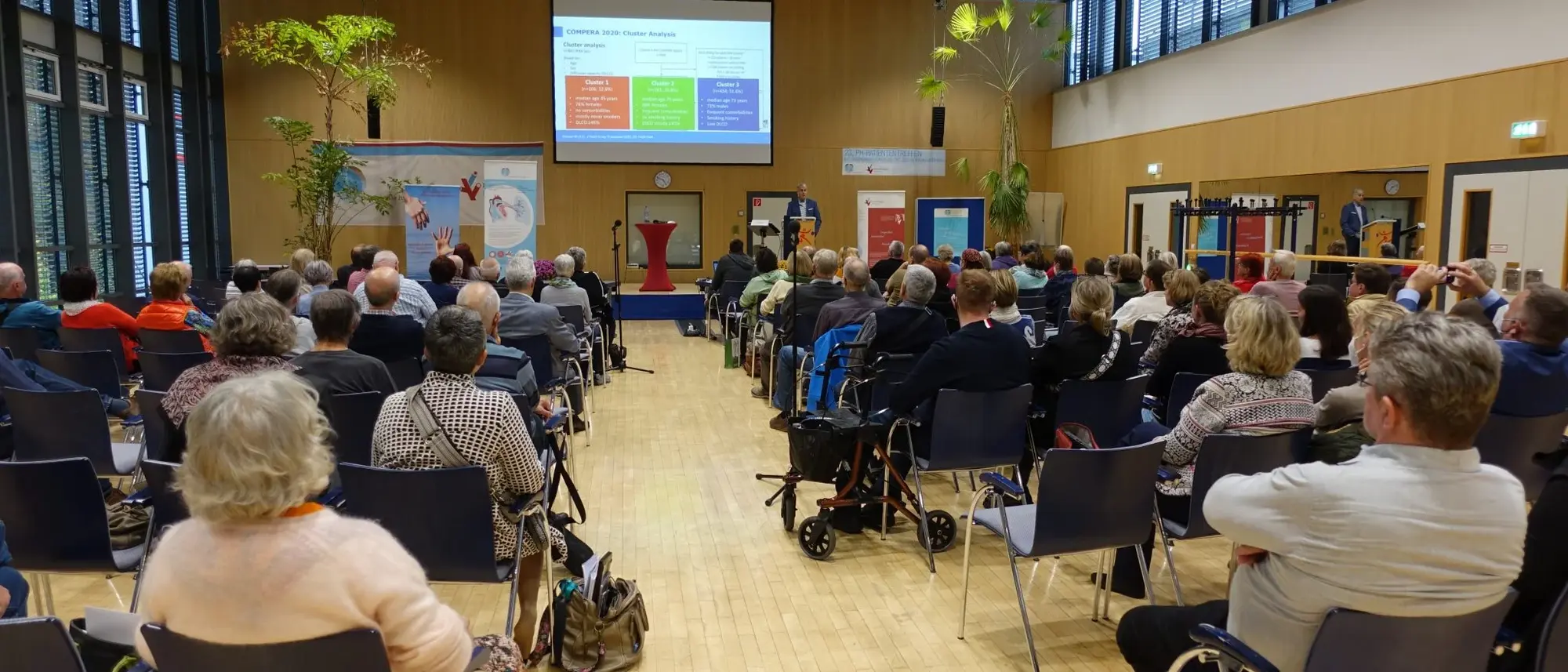 Das Bild zeigt ein PH-Patiententreffen in einem großen Saal. Viele Teilnehmer sitzen auf Stühlen und blicken auf die Bühne, wo ein Redner eine Präsentation hält. An der Wand hängt eine Leinwand, auf der eine Präsentationsfolie angezeigt wird. Im Raum sind Informationsposter und Pflanzen zu sehen, die eine angenehme Atmosphäre schaffen.