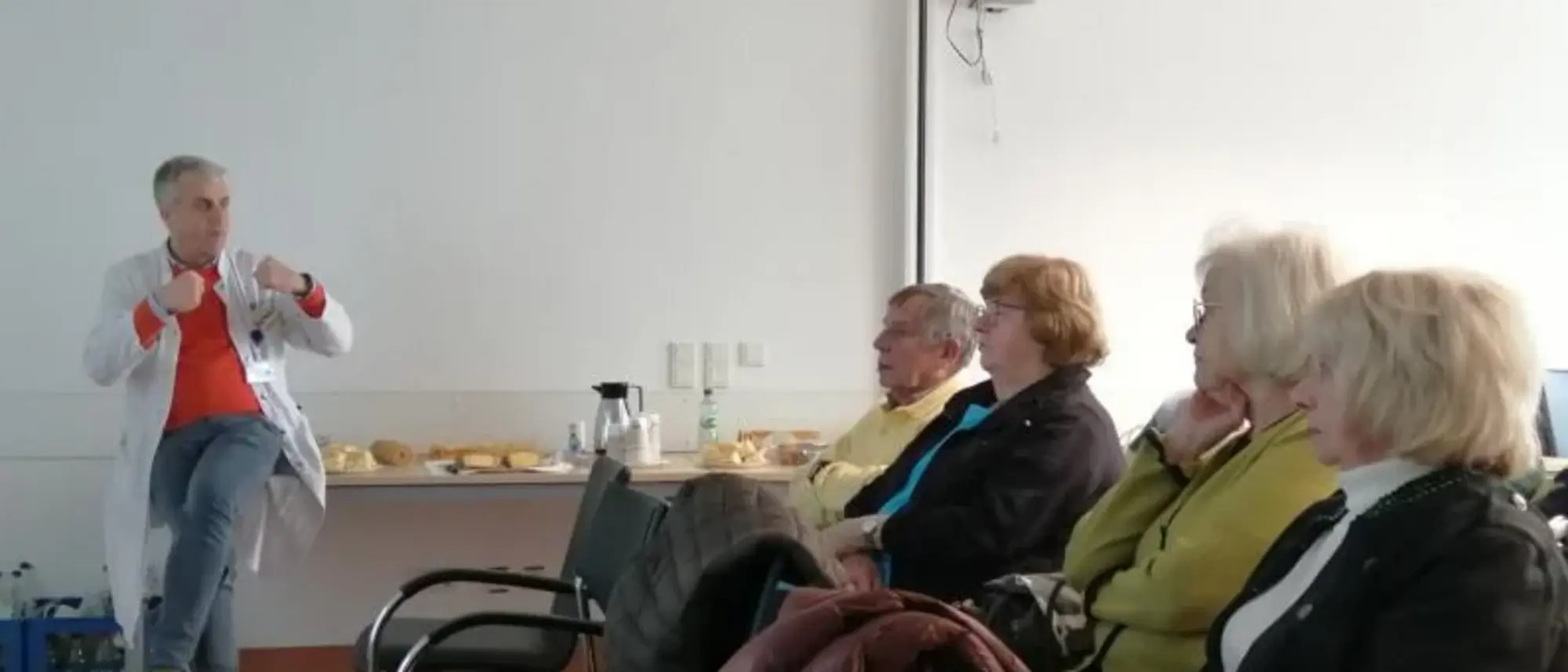 Ein Mann in einem weißen Kittel spricht zu einer Gruppe älterer Menschen, die in einem Raum auf Stühlen sitzen und aufmerksam zuhören. Im Hintergrund ist ein Tisch mit Snacks und Getränken zu sehen.