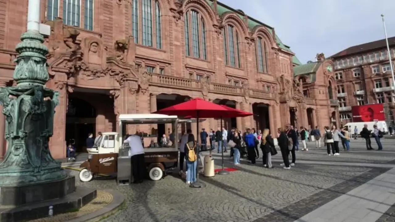 Eine historische, imposante Fassade eines Gebäudes aus rotem Sandstein, vermutlich ein Bahnhof oder ein öffentliches Gebäude. Vor dem Gebäude versammeln sich Menschen auf einem gepflasterten Platz. Ein kleiner Kaffeestand mit einem roten Sonnenschirm steht im Vordergrund, umgeben von Personen, die Getränke bestellen. 